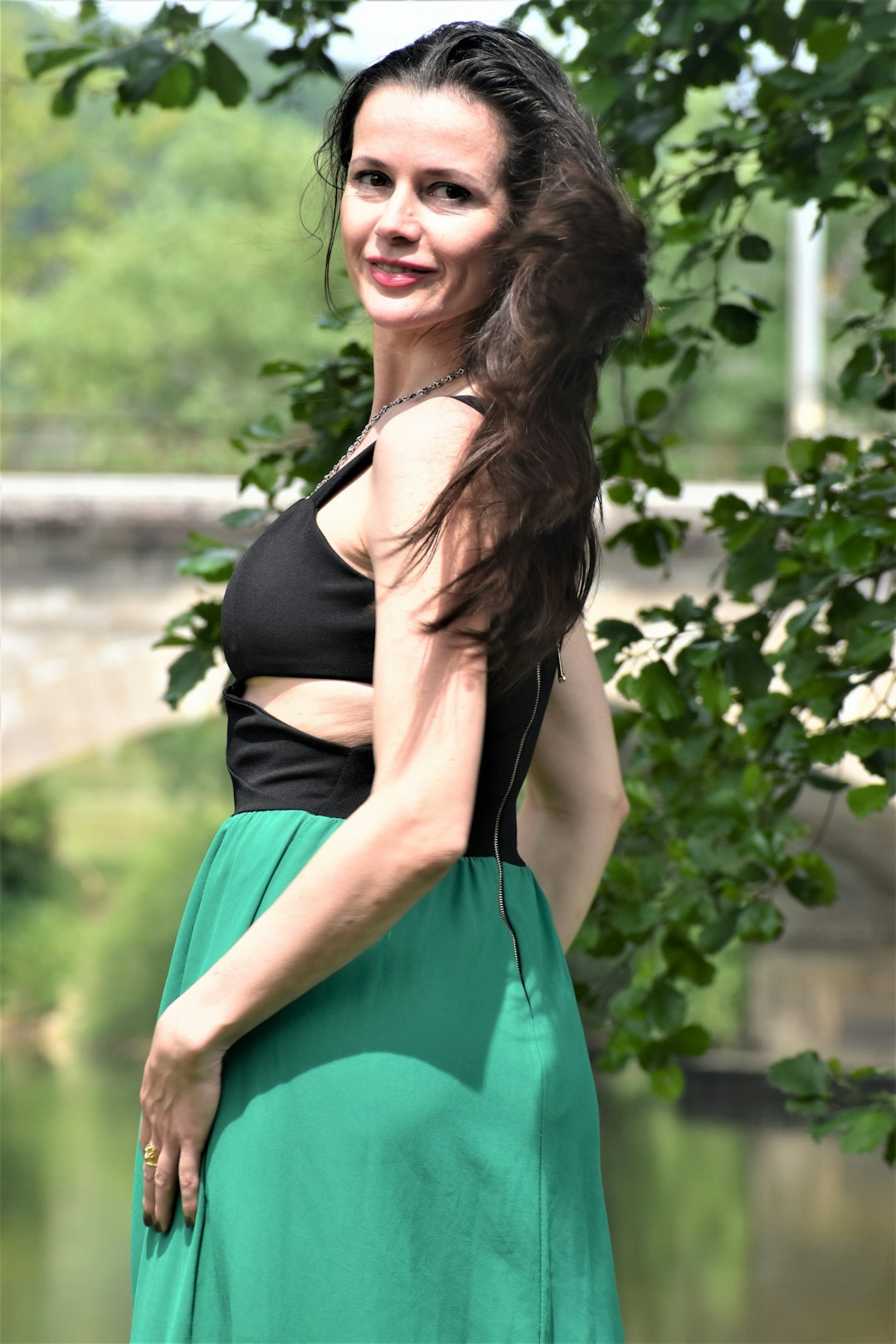 Woman in a black and green dress posing outdoors, surrounded by greenery, with flowing hair and a bridge in the background, reflecting a carefree lifestyle.